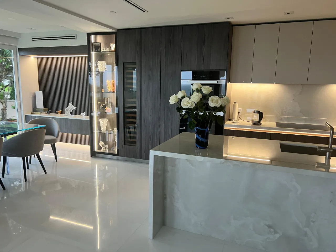 Lit glass display cabinet of decorative objects beside dark wood-grain tall cabinets with a wine column, and a veined-stone waterfall island
