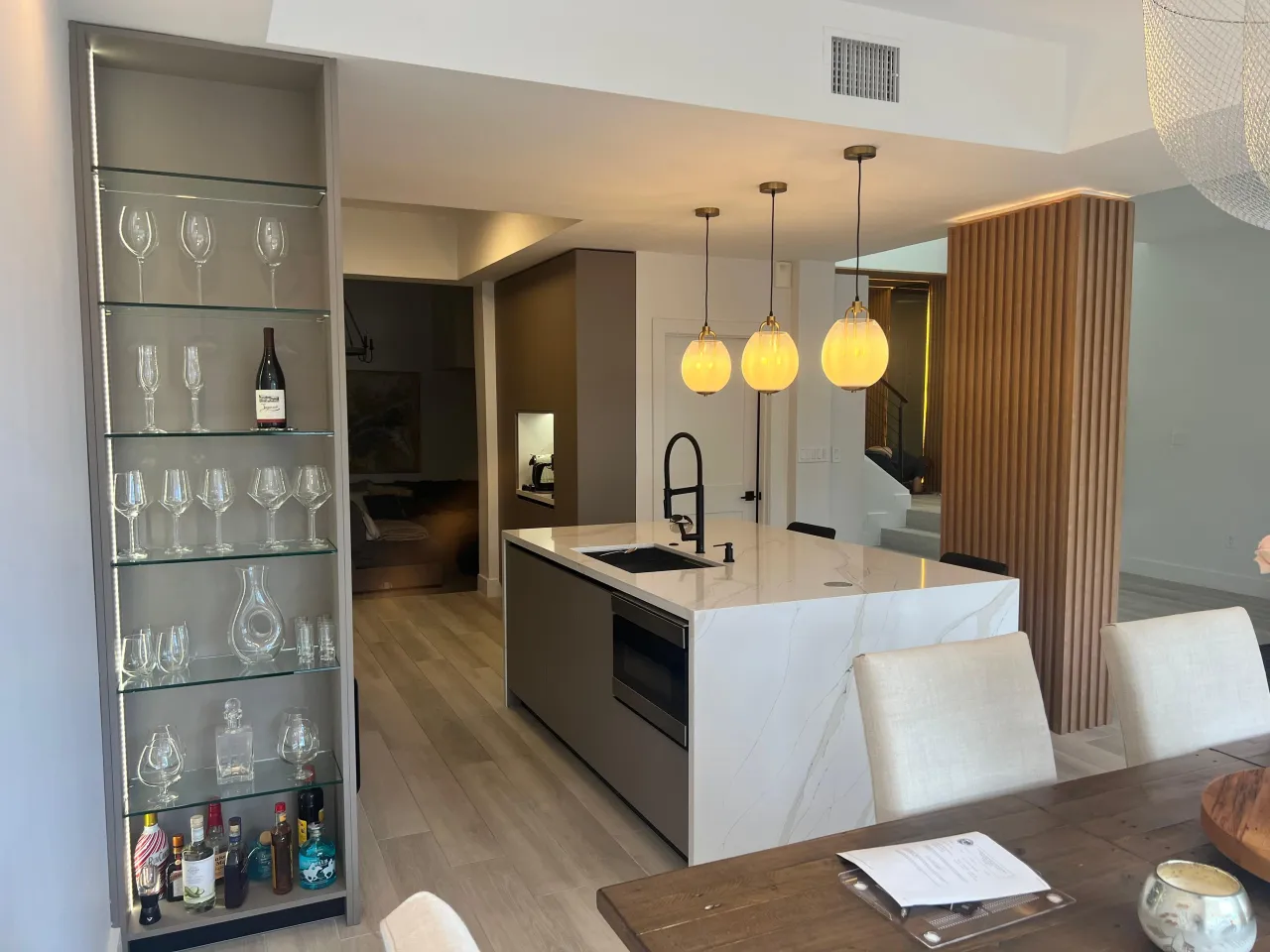 Kitchen with lit glass display shelving of glassware and bottles, a veined-stone waterfall island with a built-in oven, and three globe pendants