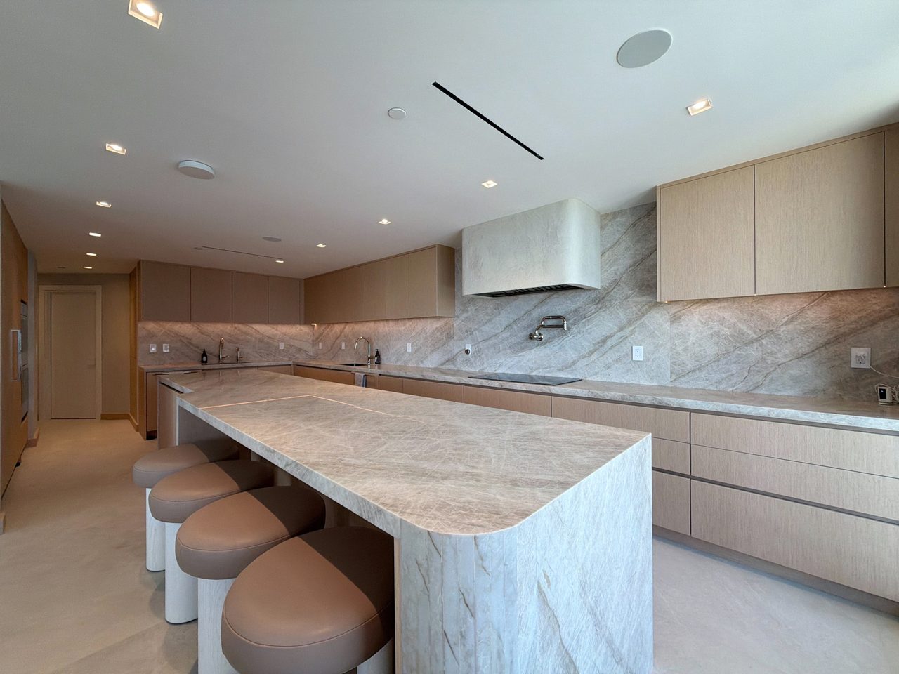 Long veined-stone waterfall island with leather-topped stools, pale wood veneer cabinetry, a wall-mounted pot filler, and an induction cooktop