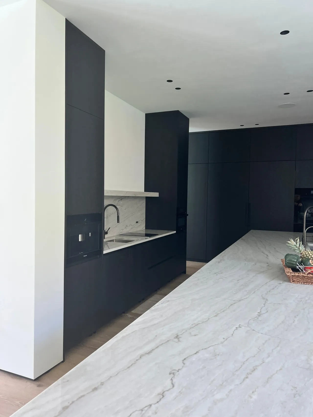 Matte black kitchen sink and cooktop area with a stone backsplash, a floating shelf, black cabinetry, and a veined-stone island counter