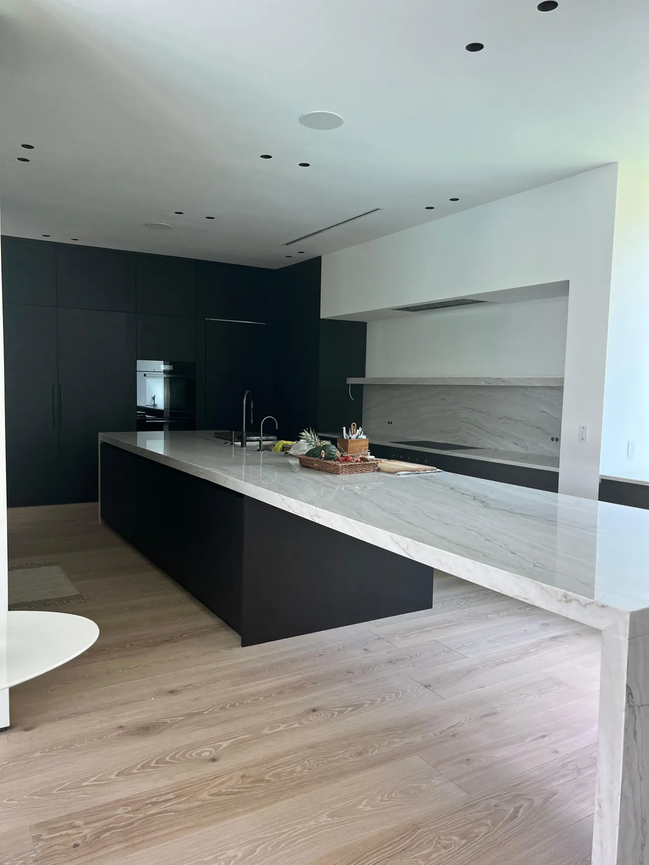 Matte black kitchen with a long veined-stone waterfall island, black tall pantry cabinetry, and a floating shelf in the backsplash niche
