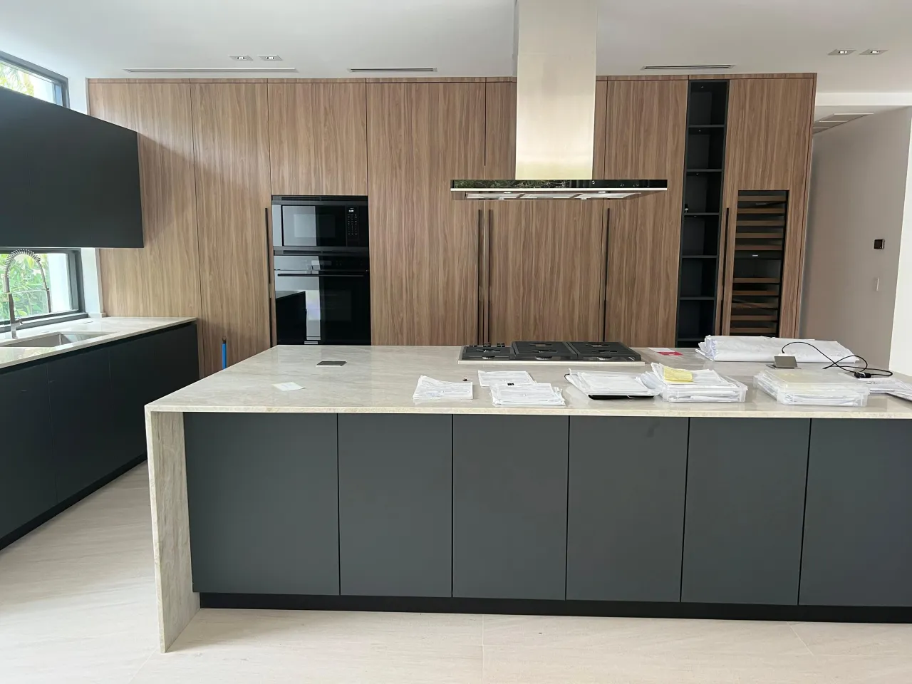 Kitchen mid-installation with wood-grain tall cabinets, a dark gray island under a light stone top with a gas cooktop, and a stainless island hood