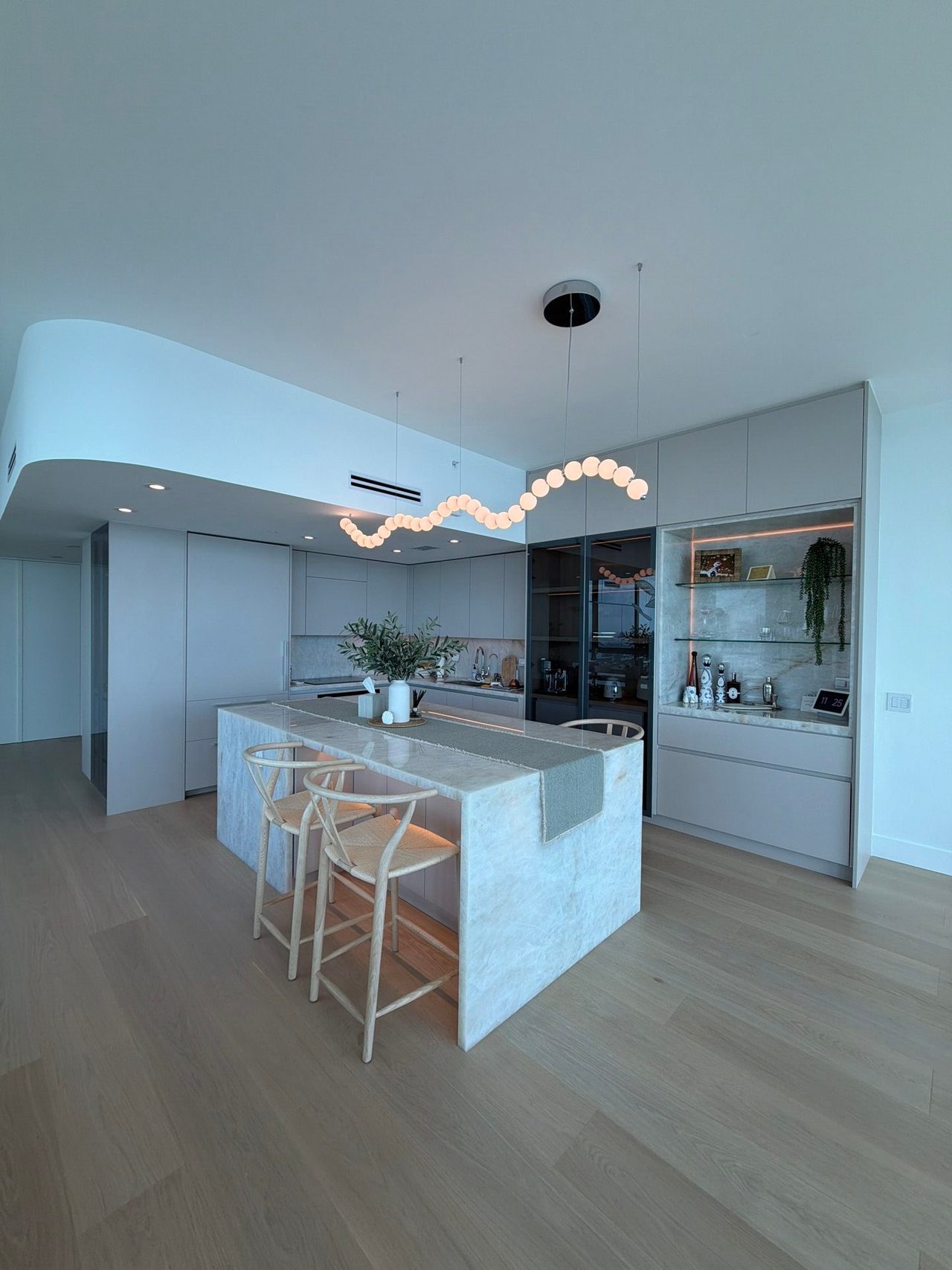 Open-concept space with a veined-stone-top island, rattan barstools, a curved white soffit, and a string-of-bubbles pendant light over light wood flooring