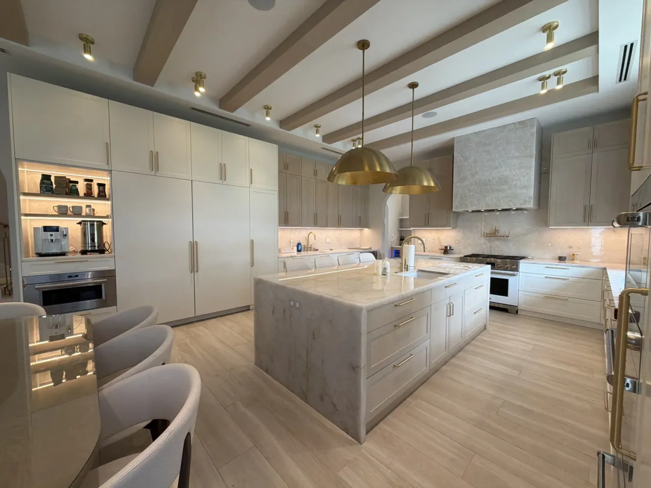 Open kitchen with cream flat-panel cabinets, a white veined-stone waterfall island, two gold-tone dome pendants, and a melamine-clad beam ceiling