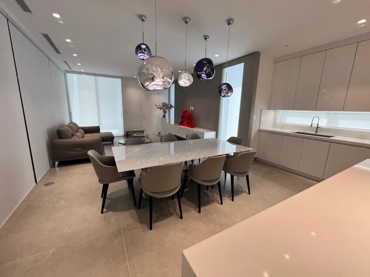 Open kitchen and dining space with five iridescent metallic globe pendants over a veined-stone table, pale flat-front cabinetry and a sink run