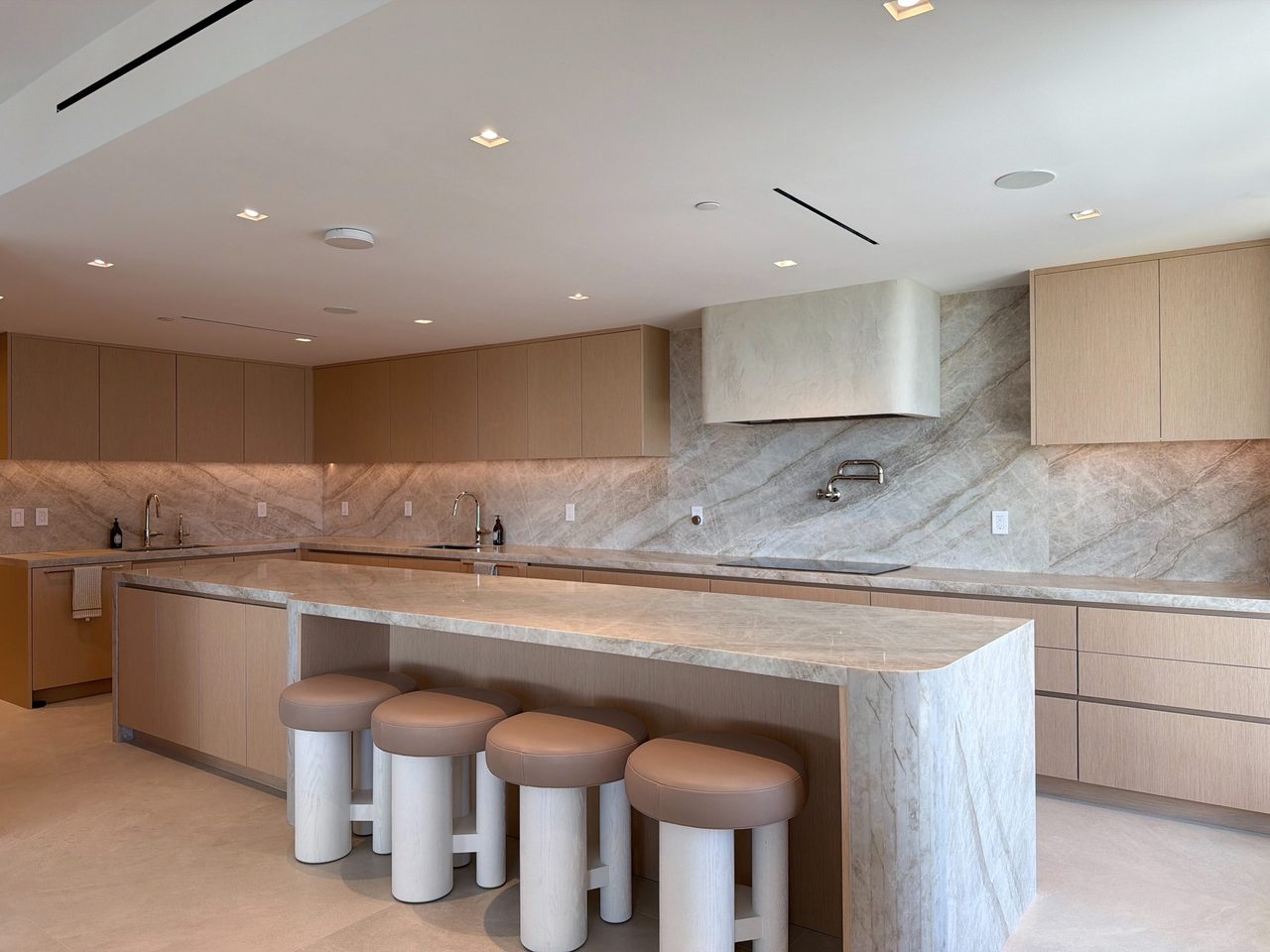 Pale custom-stained wood veneer kitchen with a long veined-stone waterfall island, leather-topped stools, and a stone-wrapped hood over a slab backsplash