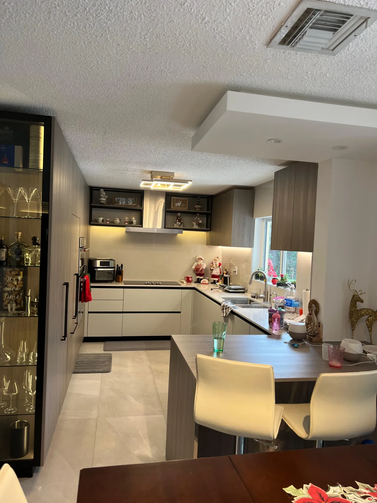 Kitchen with pale flat-front cabinets, a lit glass display cabinet of glassware, a black cooktop under a stainless hood, and a wood-grain peninsula