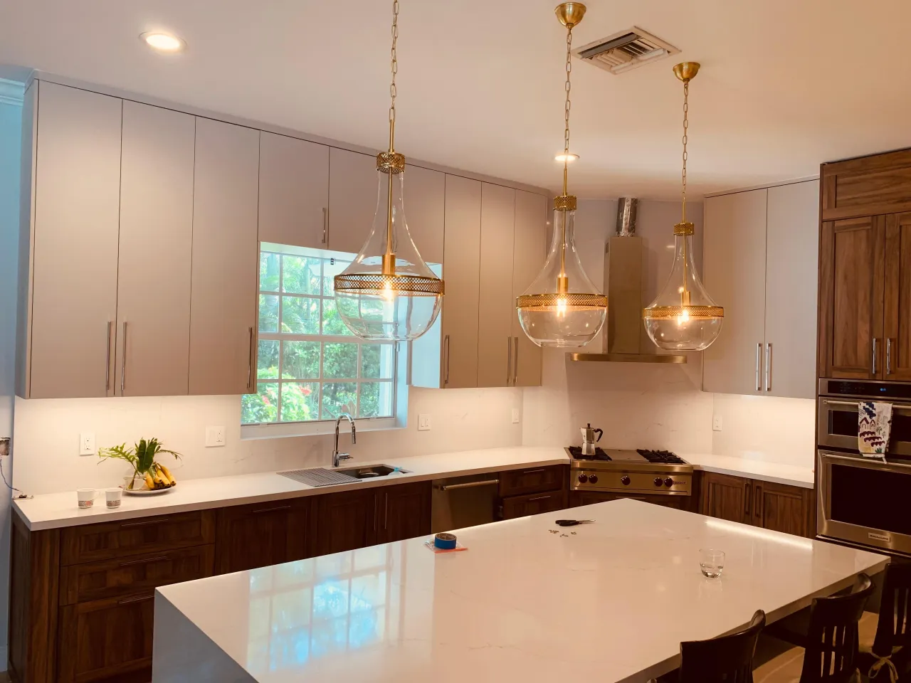 Kitchen with pale gray uppers and dark wood-grain base cabinets, three glass and gold-tone pendants over a white island, a stainless range and hood