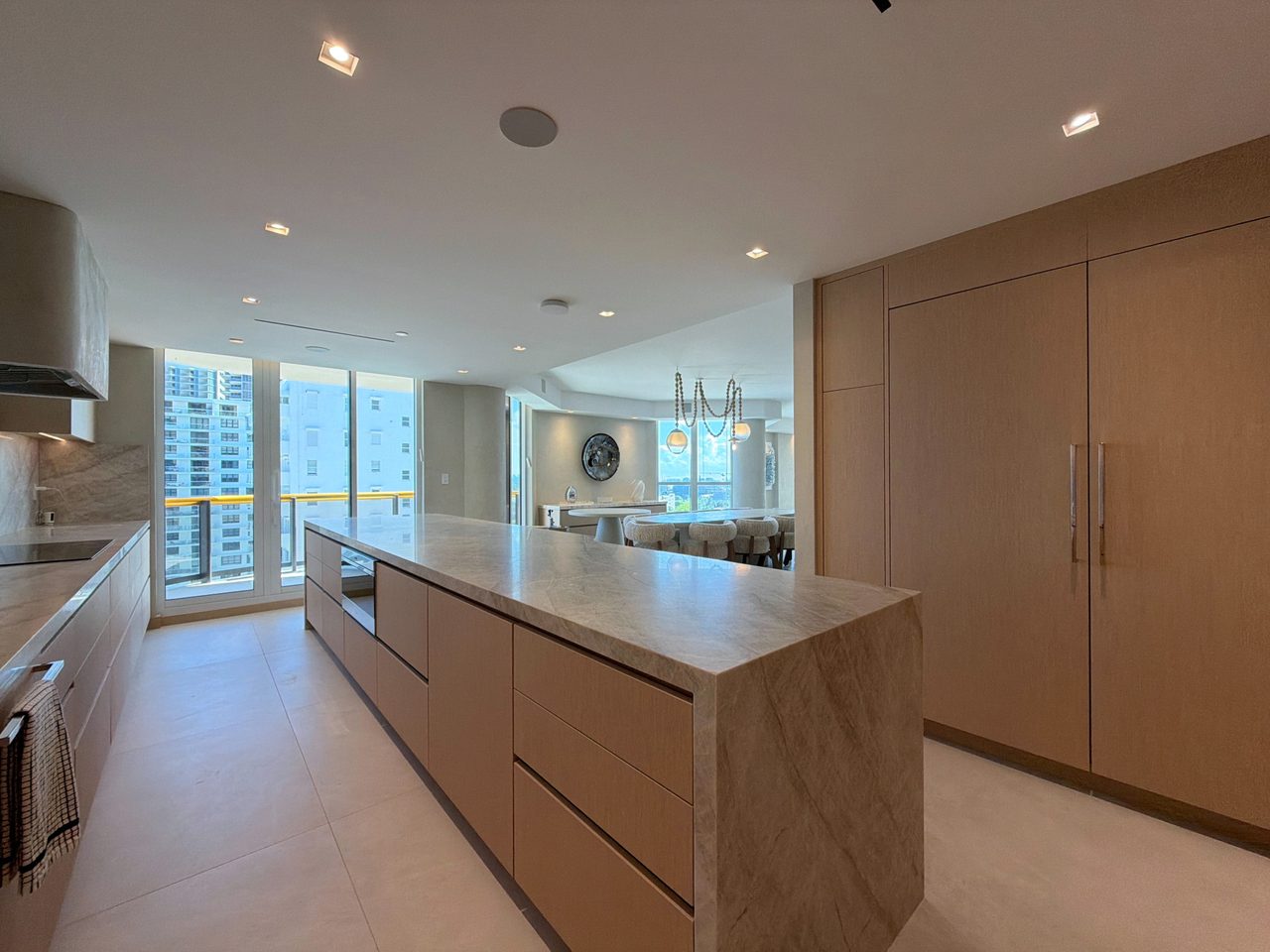 Pale custom-stained wood veneer kitchen with a veined-stone island and a tall integrated refrigerator wall beside floor-to-ceiling windows