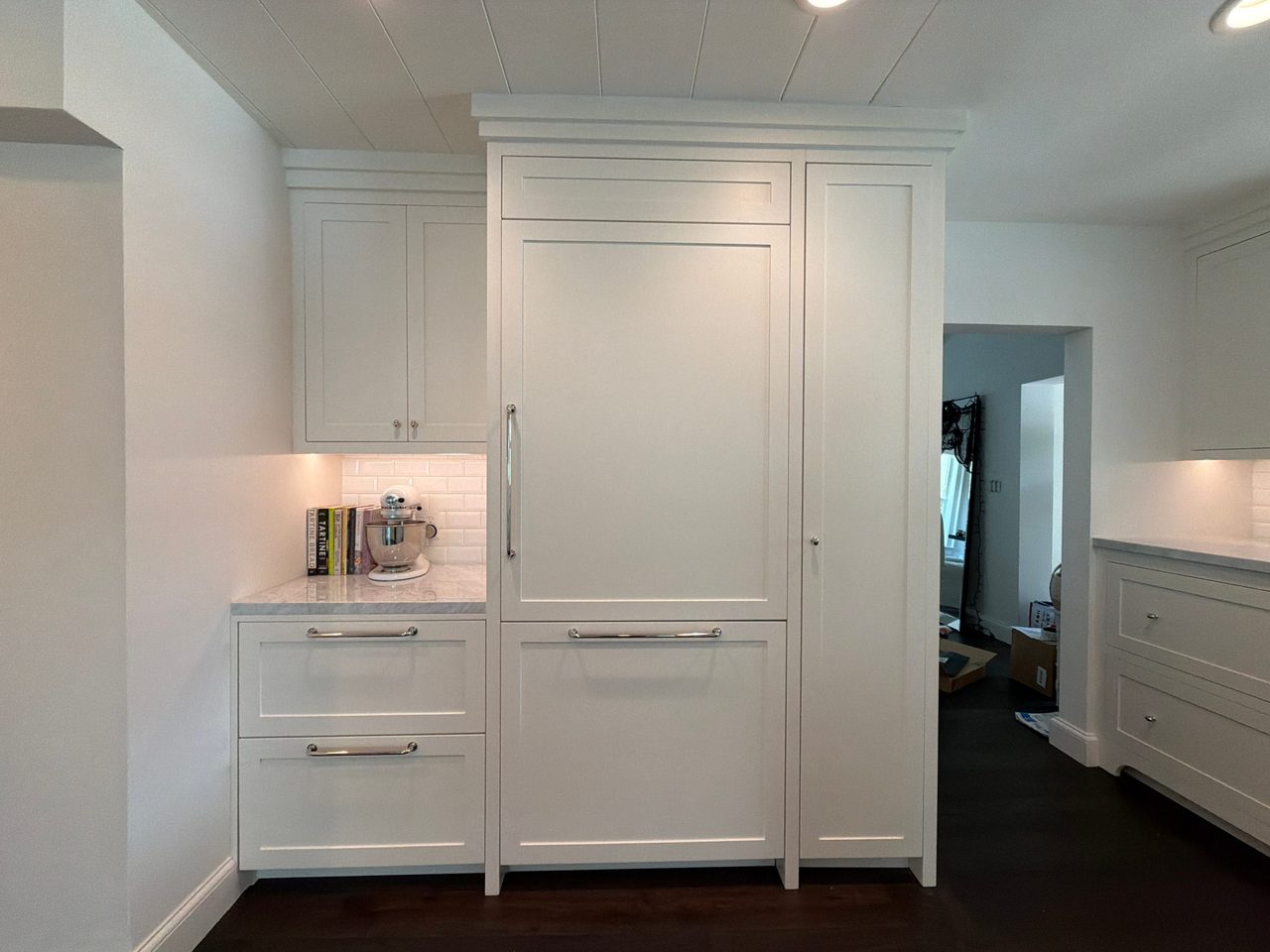 Paneled built-in refrigerator with a full-height pantry cabinet, white shaker upper cabinets, drawer bases, and chrome bar pulls on a dark wood floor