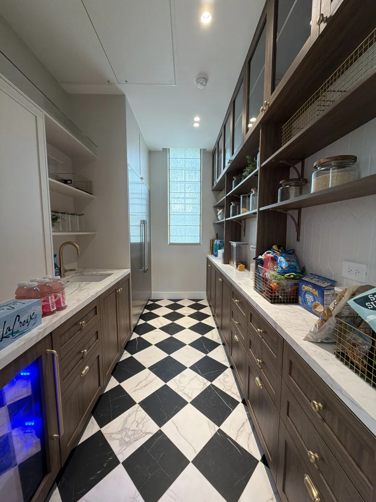 Pantry aisle with dark wood-grain cabinets and open shelving on one side, a sink run with a blue-lit beverage fridge opposite, and a checkerboard floor