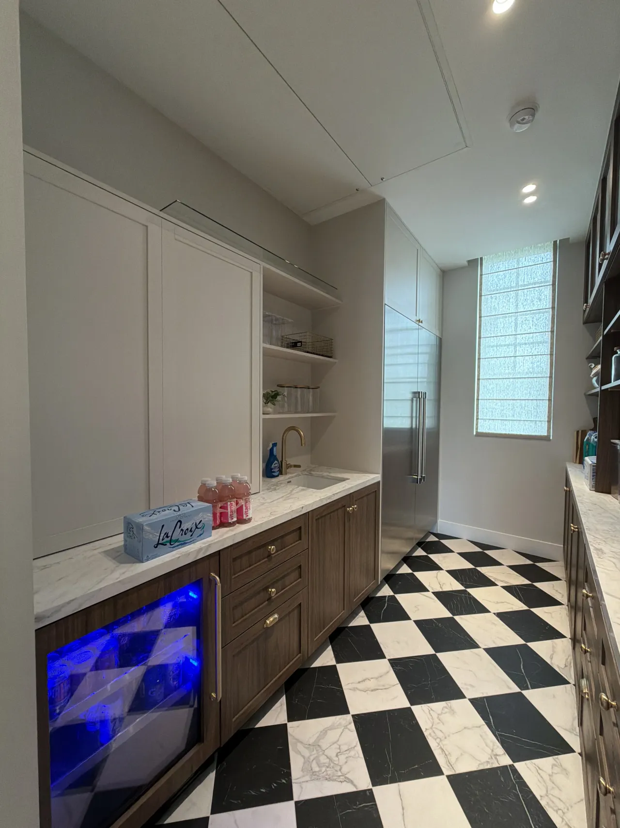 Pantry end wall with white upper cabinets, a glass shelf, a sink with gold-tone faucet, a blue-lit beverage fridge, and a checkerboard floor