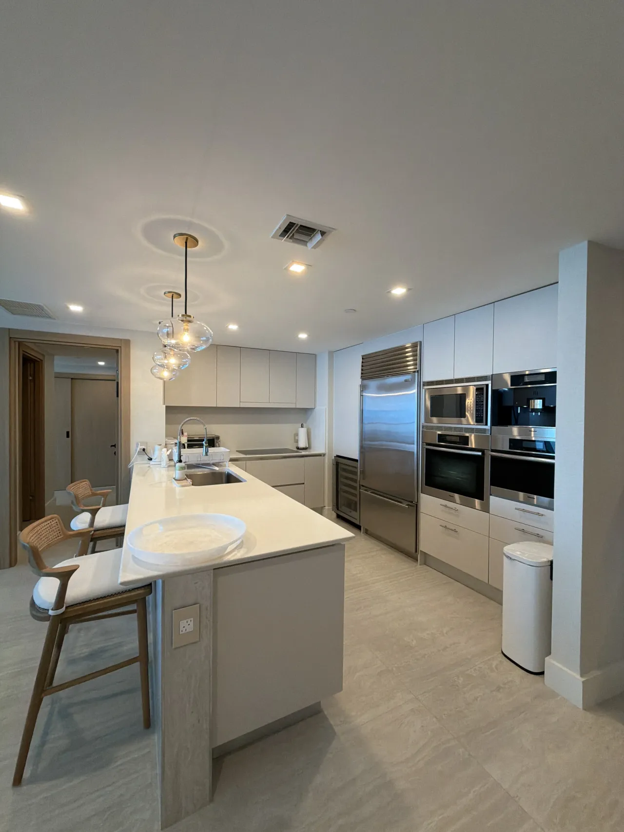 Kitchen peninsula with a white counter and two woven-back barstools, glass globe pendants, a stainless fridge, and stacked wall ovens