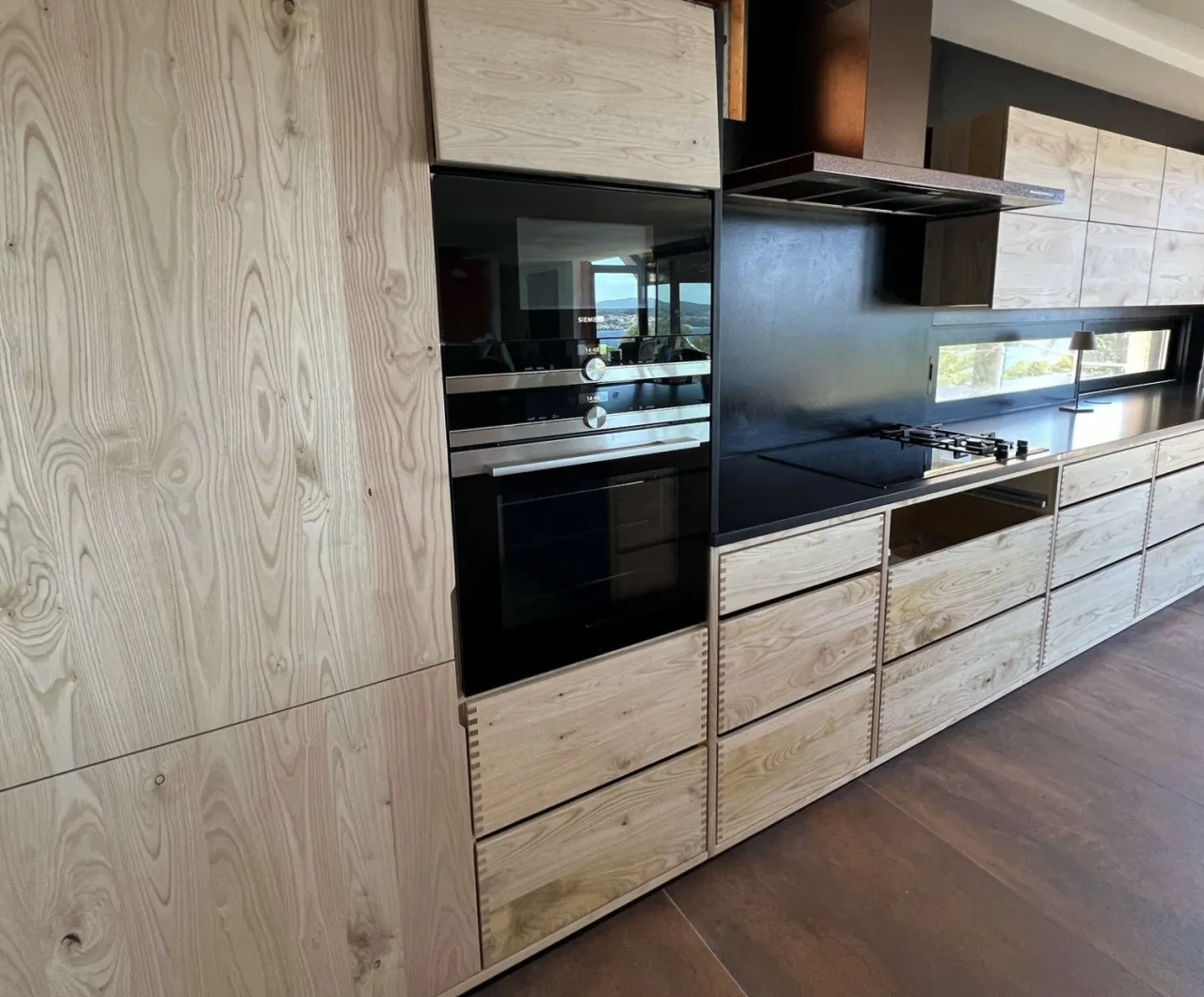 Run of pale raw-wood cabinetry with dovetailed drawers, black built-in ovens and cooktop against a black backsplash and counter, over a dark wood floor