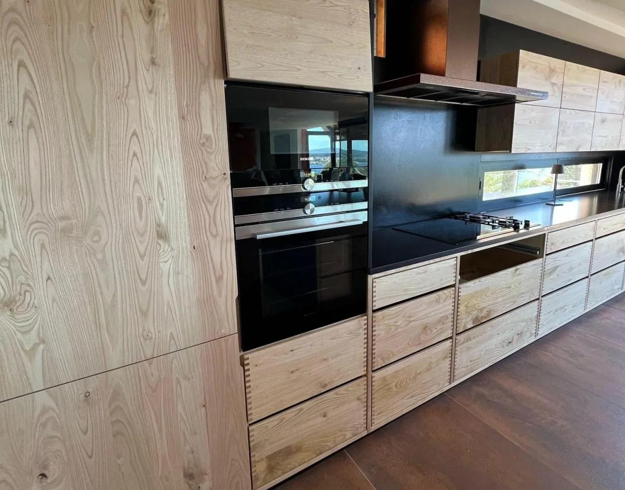 Rustic light wood kitchen with a black glass backsplash and cooktop, dovetail-joint drawer fronts, and a black integrated oven