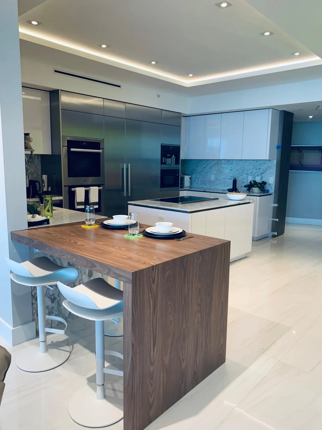 Stainless appliance wall kitchen with a dark wood breakfast bar/table island, a veined-stone backsplash, and white glossy cabinetry