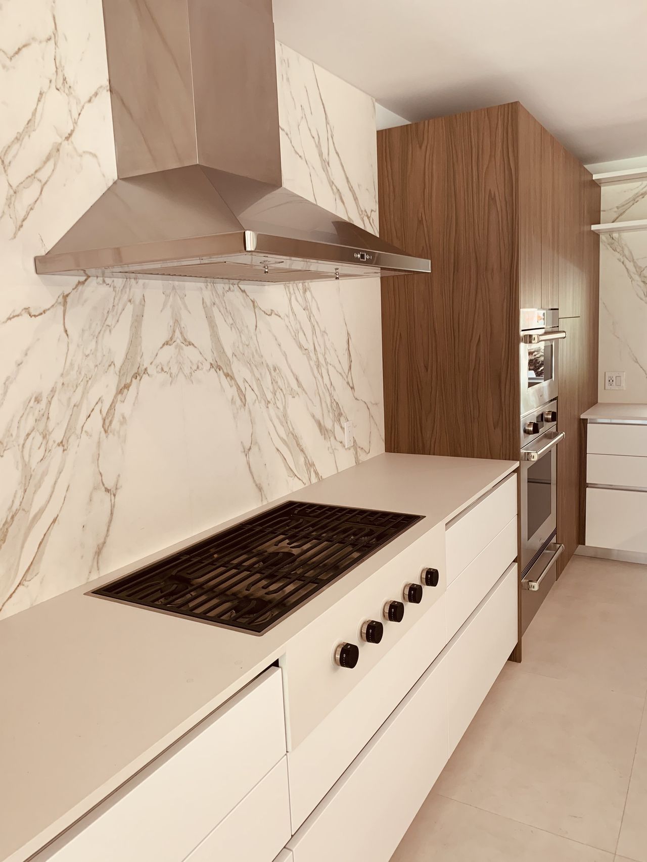 Stainless range hood, veined-stone slab backsplash, and a gas cooktop set into a white countertop with dark wood cabinetry beside it