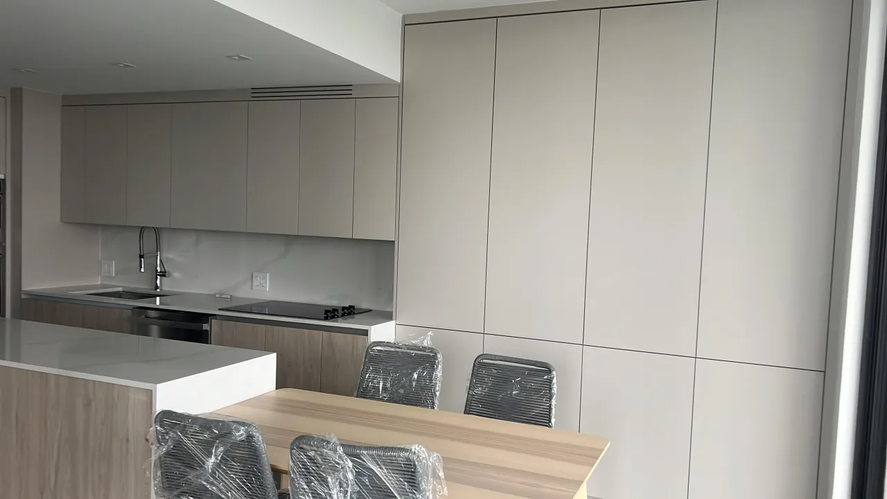 Taupe kitchen cabinetry and a tall cabinet wall, with a white waterfall island counter and an attached light wood-grain dining table with wrapped chairs