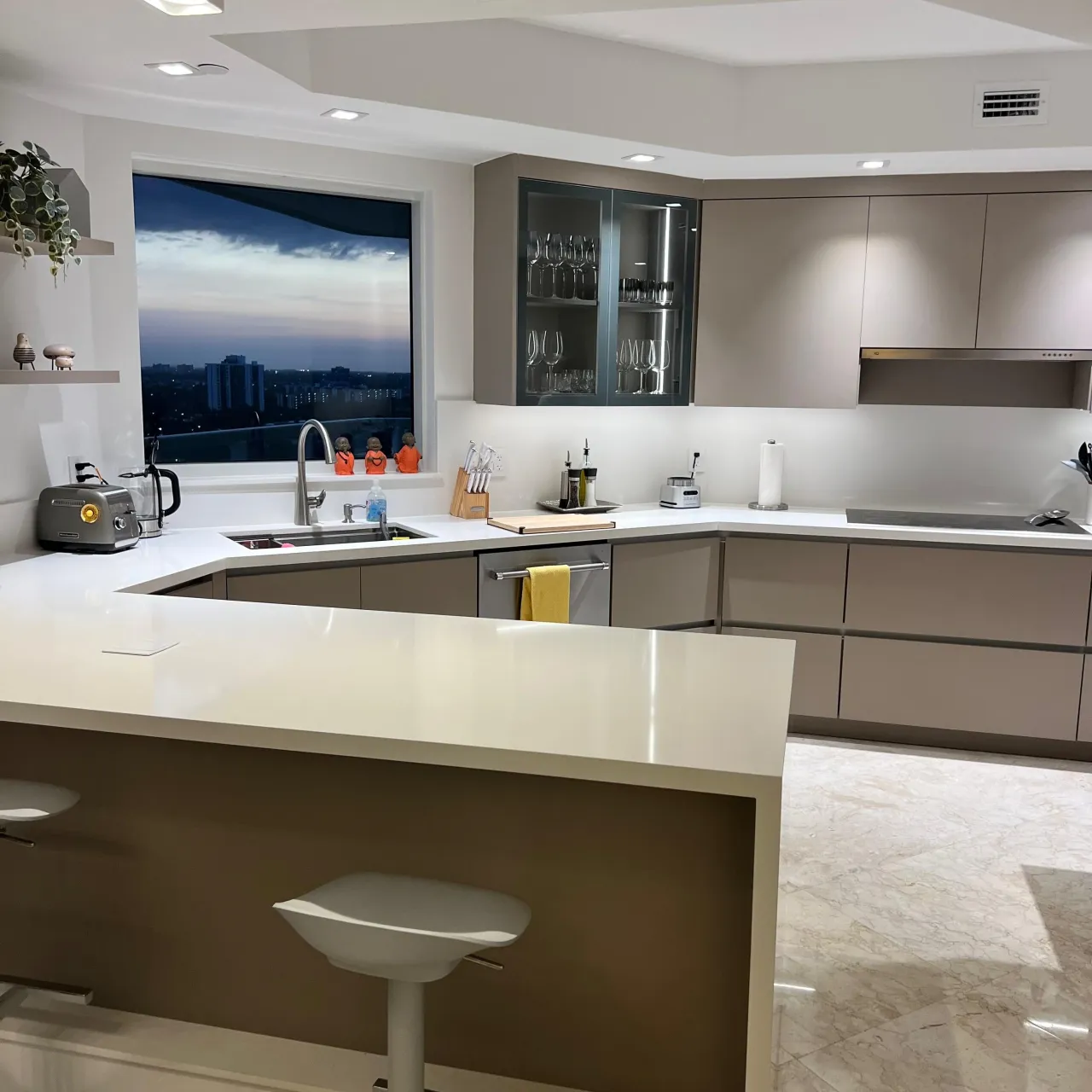 Taupe/gray kitchen with a glass display cabinet, white stone counters, a night skyline view through the window, and a U-shaped peninsula