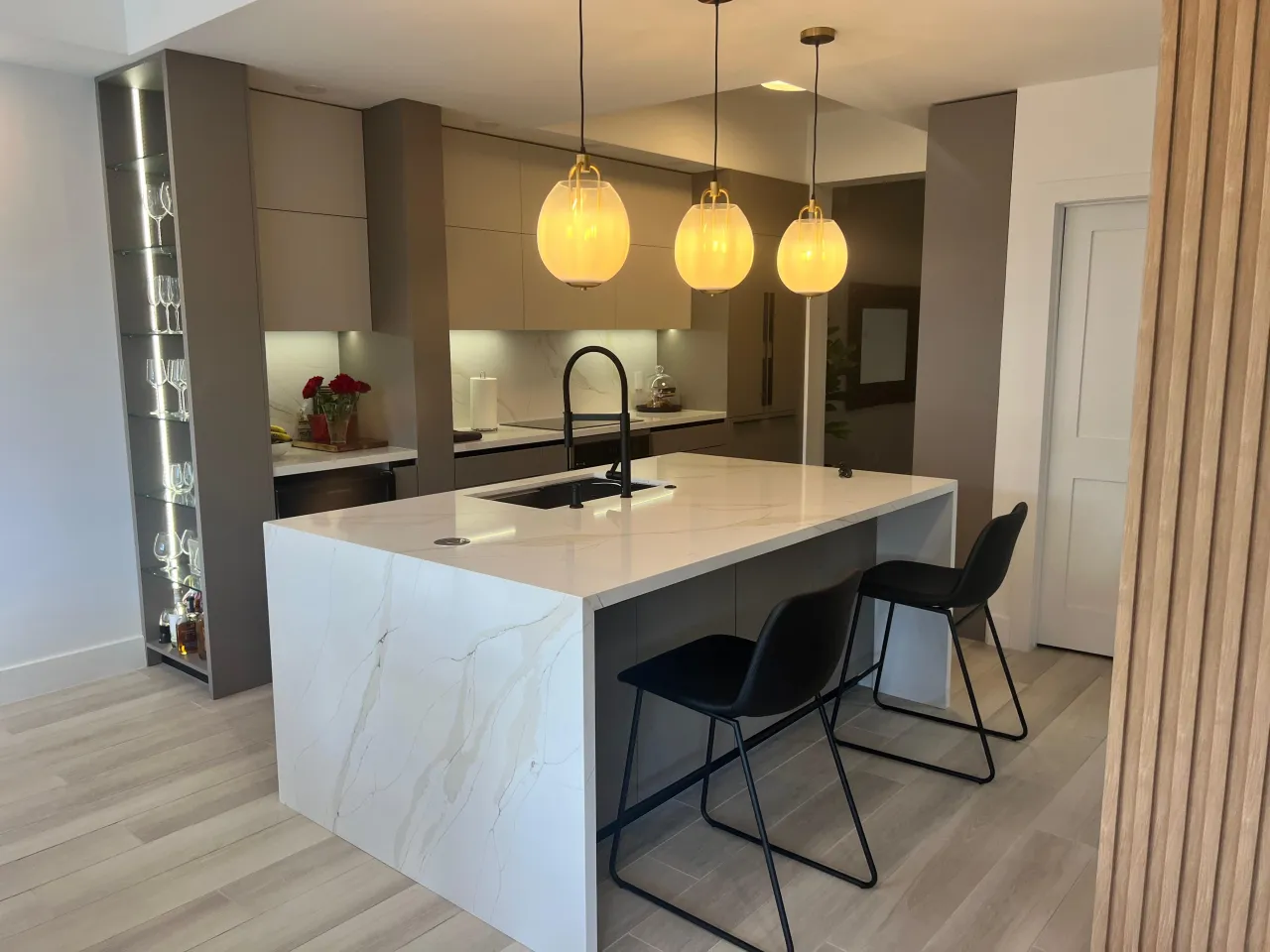 Taupe kitchen with a veined-stone waterfall island, three amber glass globe pendant lights, and a glass wine display shelf