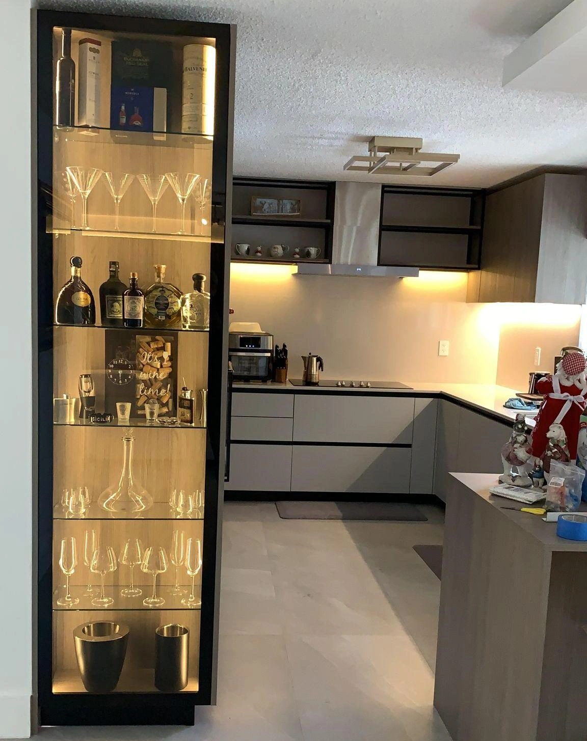Two-tone gray/glass kitchen with an illuminated glass wine display cabinet, wood-toned cabinets, and a stainless range hood