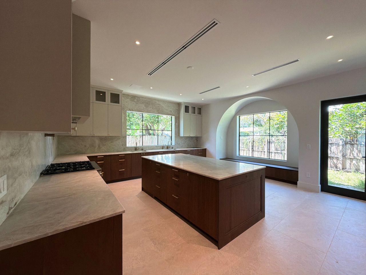 Open kitchen with two-tone melamine cabinetry, dark wood-grain and cream, a veined-stone island, a gas cooktop, and an arched window over a built-in bench