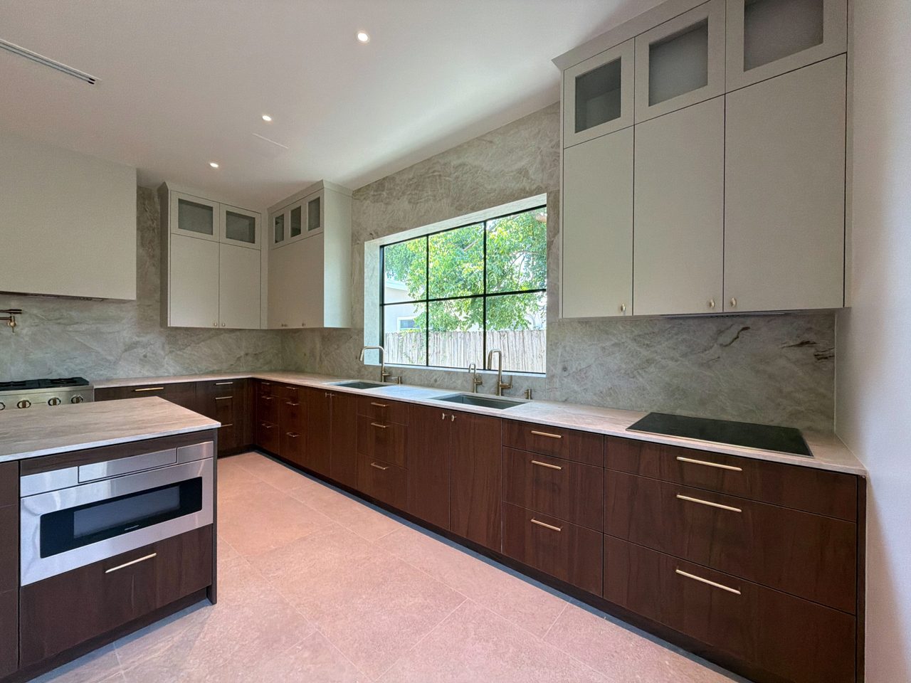 Two-tone melamine kitchen run with dark base cabinets, cream uppers, a veined-stone backsplash, double sinks below a steel window, and an induction cooktop