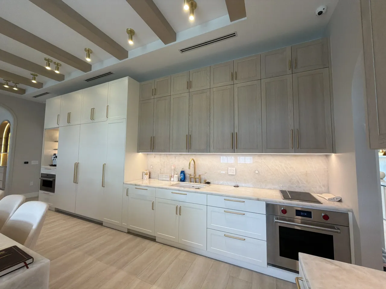 Two-tone kitchen run with taupe upper cabinets, white lower cabinets, veined-stone slab backsplash, gold-tone pulls, and a stainless oven under a cooktop