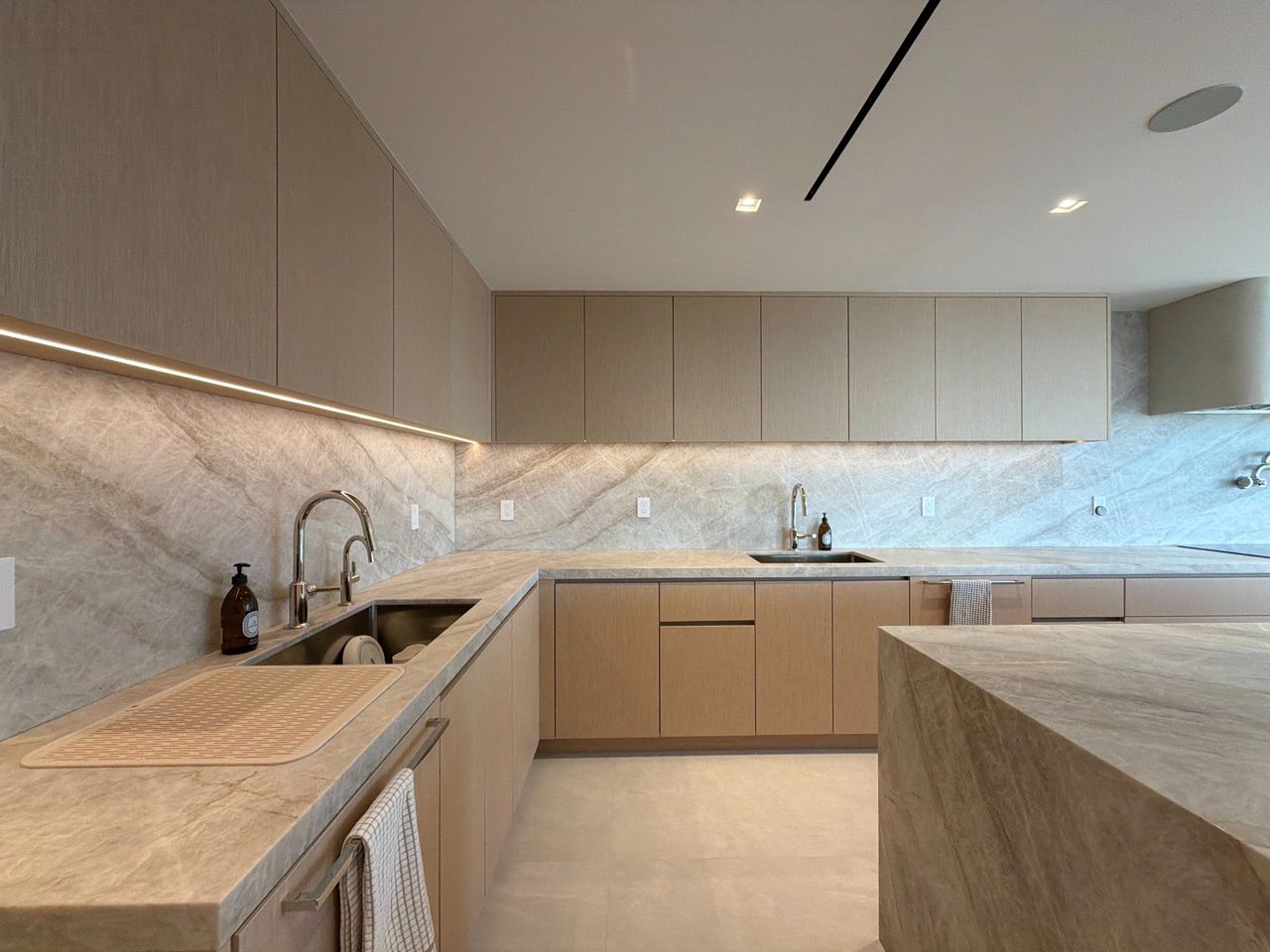 Corner of a veined-stone counter and slab backsplash with two undermount sinks, and pale custom-stained wood veneer cabinets above and below