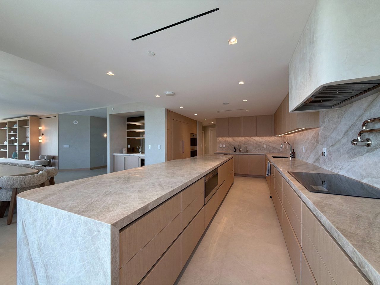 Veined-stone island with a built-in oven and pale custom-stained wood veneer cabinetry, opening to a dining area with bouclé chairs