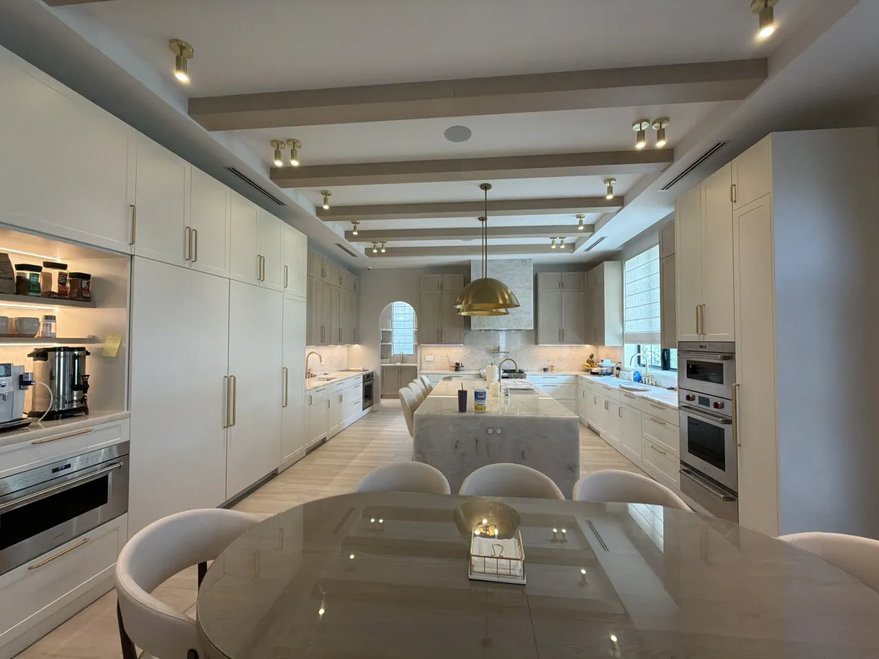 Long kitchen with a veined-stone island, cream cabinetry, a melamine-clad beam ceiling, a gold-tone pendant, and a round glossy dining table