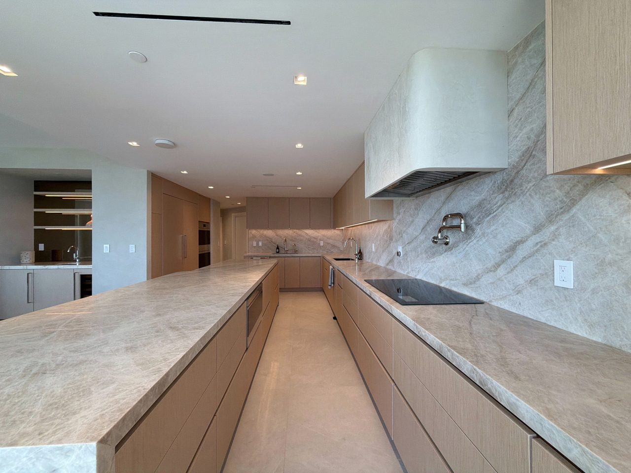 View along a veined-stone island toward the wet bar, with pale wood veneer cabinetry, a built-in island oven, and a stone-wrapped range hood