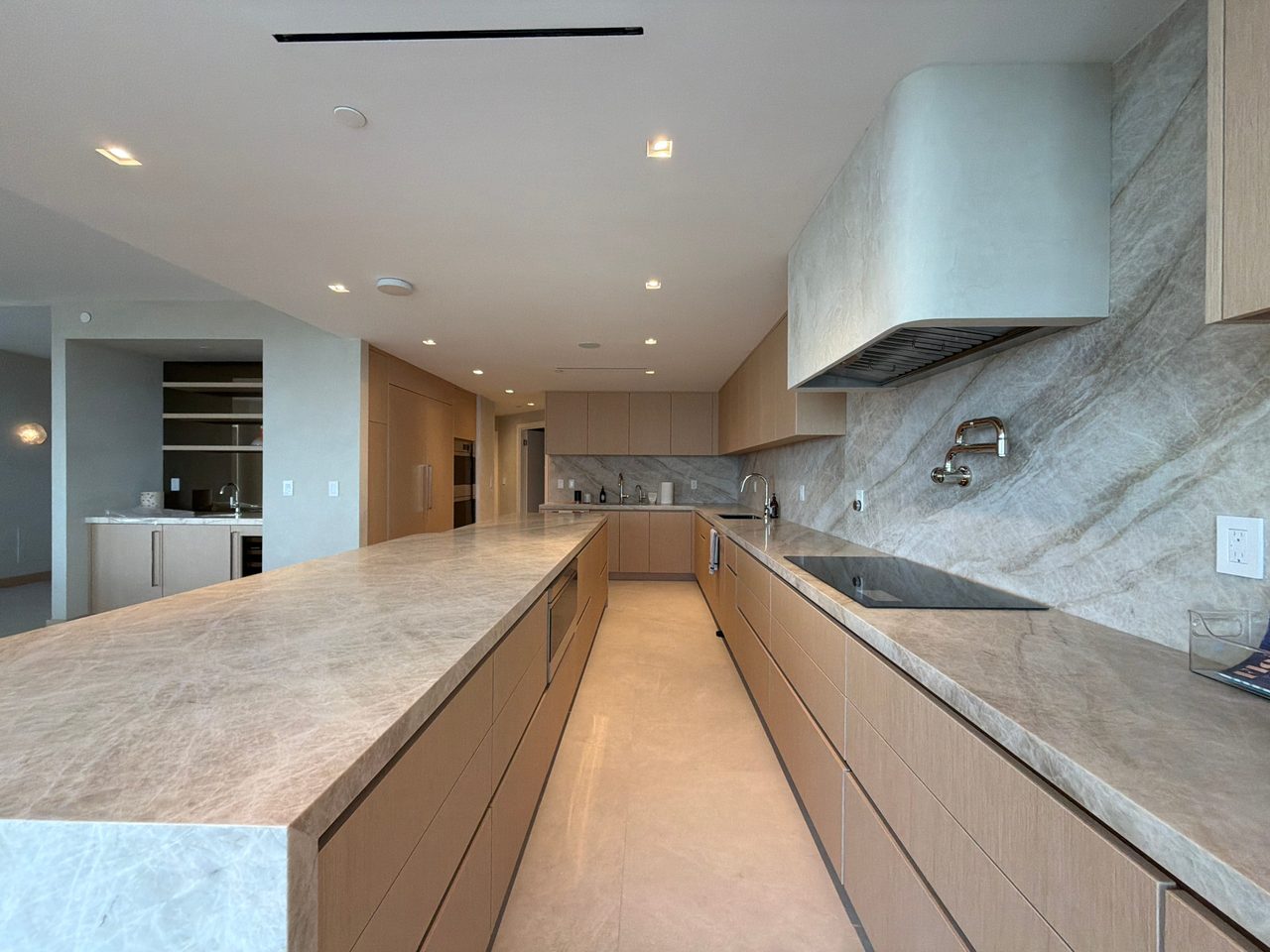 Veined-stone waterfall island beside a custom-stained wood veneer cabinet run, with a slab backsplash, wall-mounted pot filler, and induction cooktop