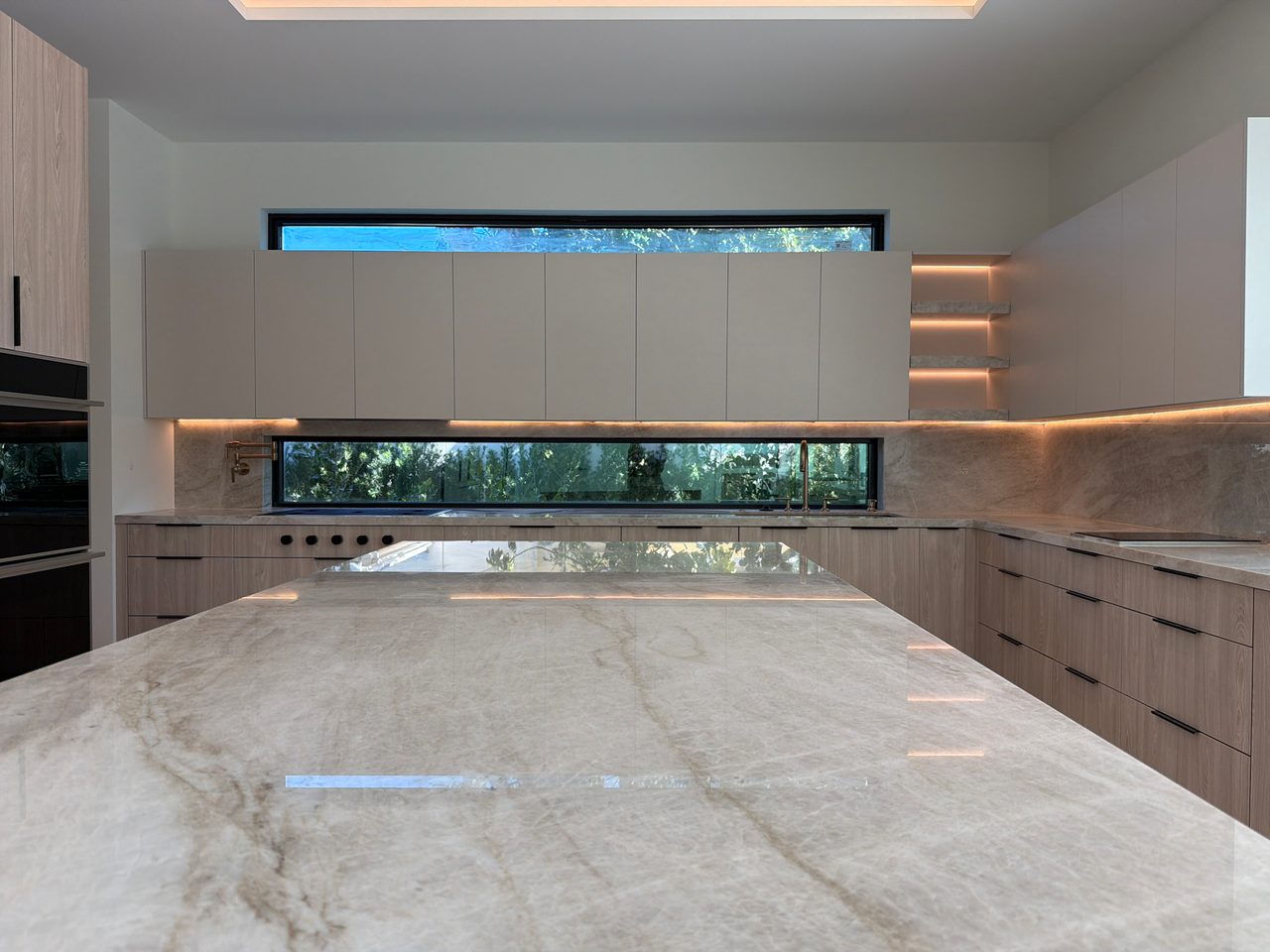 Veined-stone waterfall island countertop with light wood-grain melamine cabinetry, a narrow ribbon window over the sink, and warm LED-lit open shelving