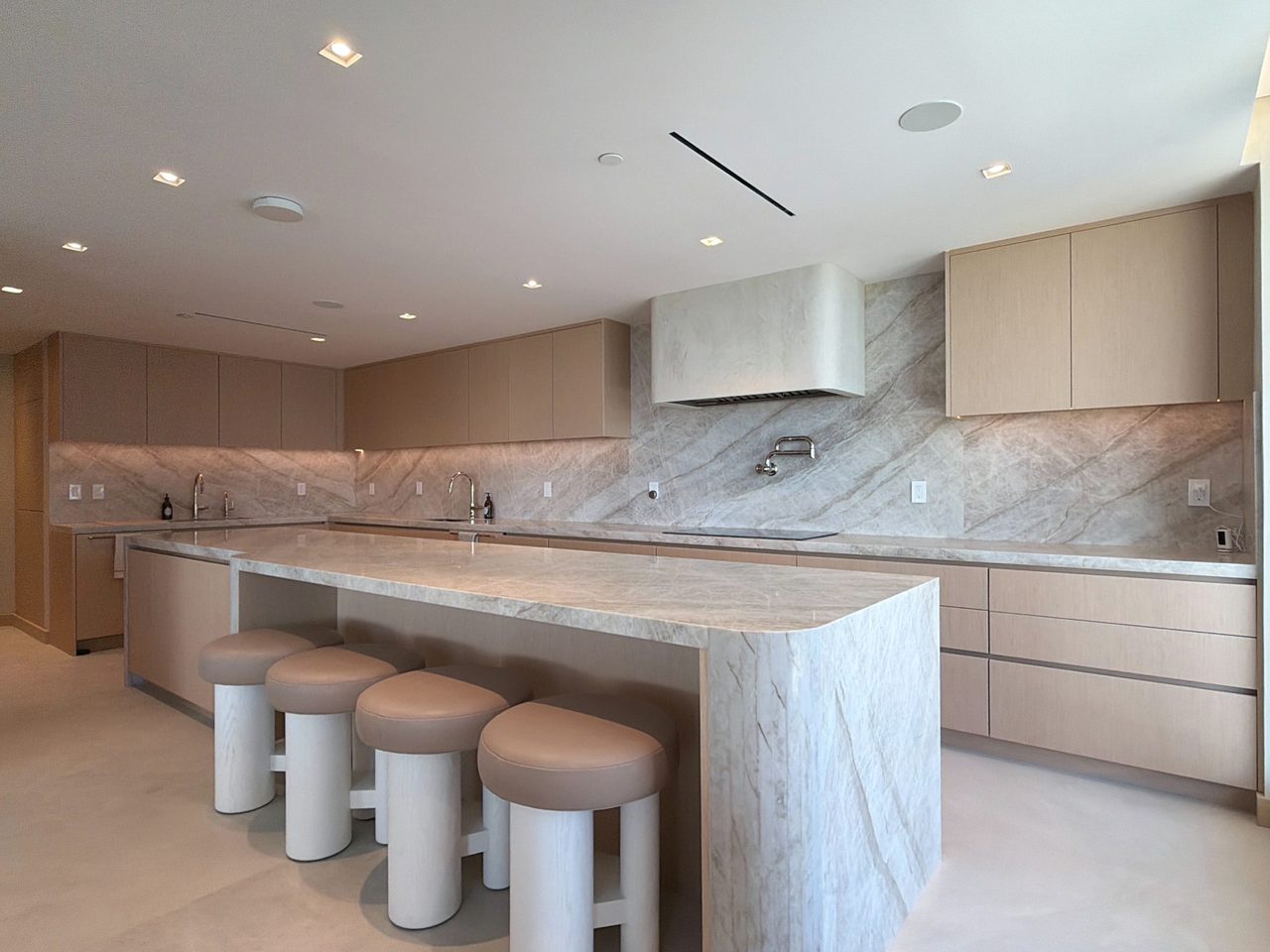 Veined-stone waterfall island and leather-topped stools in a pale custom-stained wood veneer kitchen, with an induction cooktop on the back run