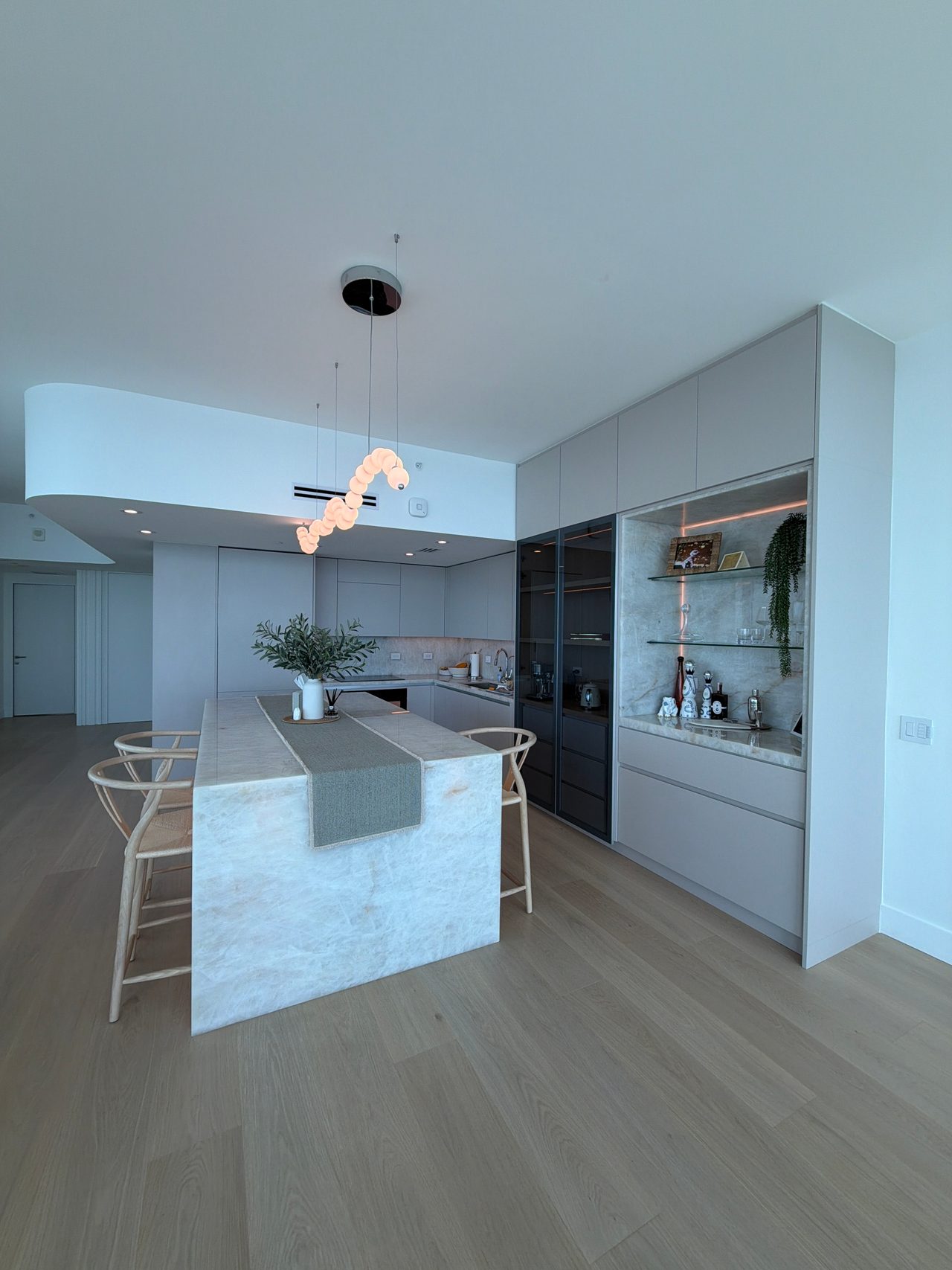 Kitchen with a veined-stone waterfall island, woven-seat stools, a wavy bubble pendant, and dark glass-front tall cabinets beside a lit display niche