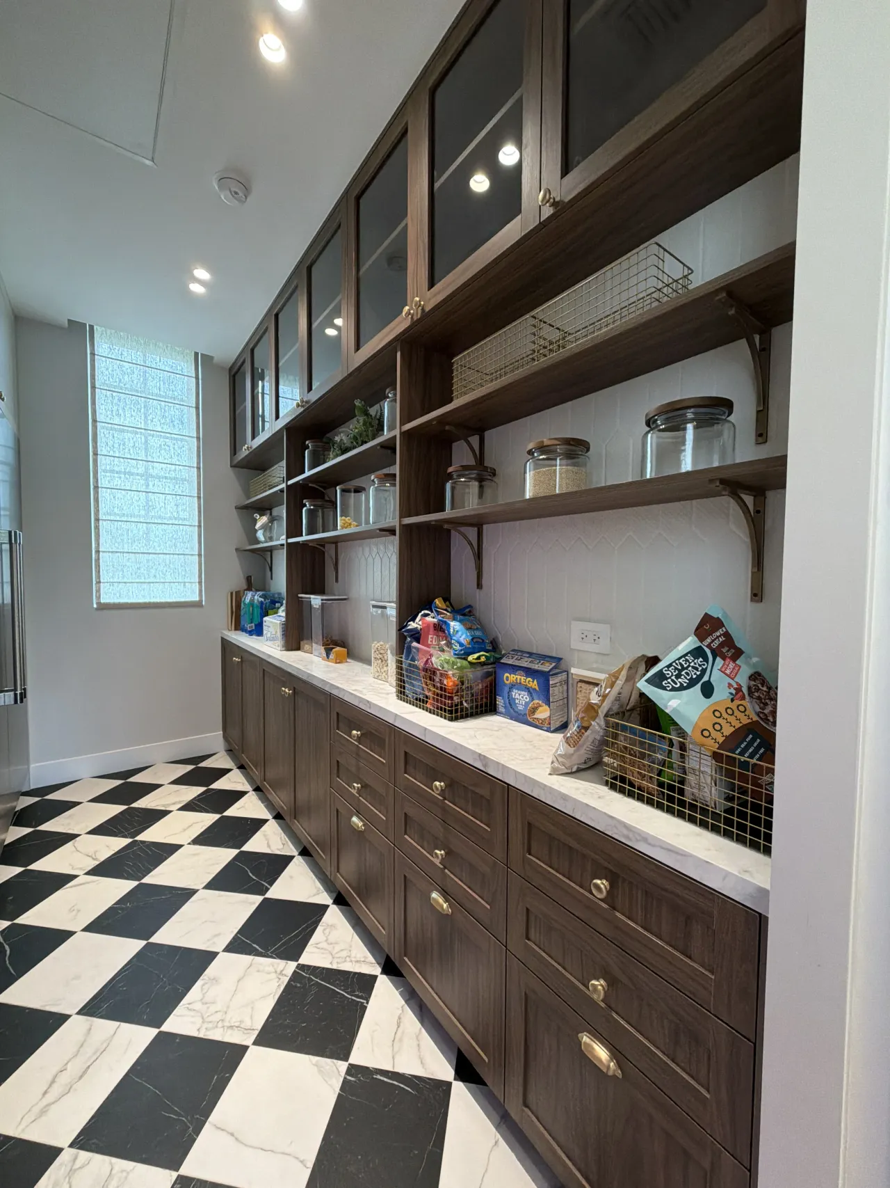 Walk-in pantry with dark wood-grain cabinets, glass-front uppers, open shelves of jars and wire baskets, a veined-stone counter, and a checkerboard floor