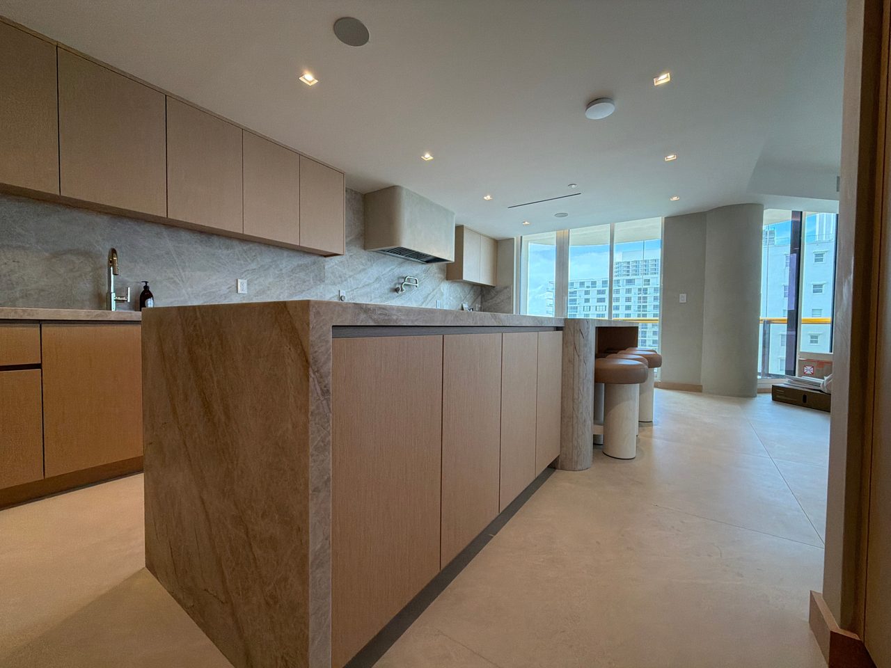 Veined-stone waterfall island with custom-stained wood veneer door fronts, opening to a living area with floor-to-ceiling windows
