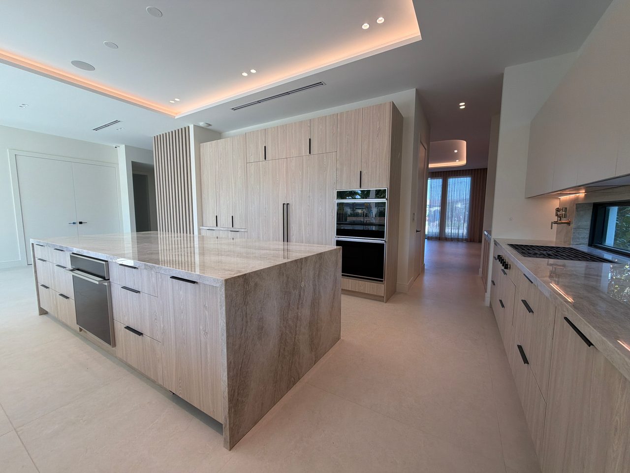 Large waterfall island in veined stone with light wood-grain melamine cabinetry, black bar pulls, and a stainless drawer appliance in the island