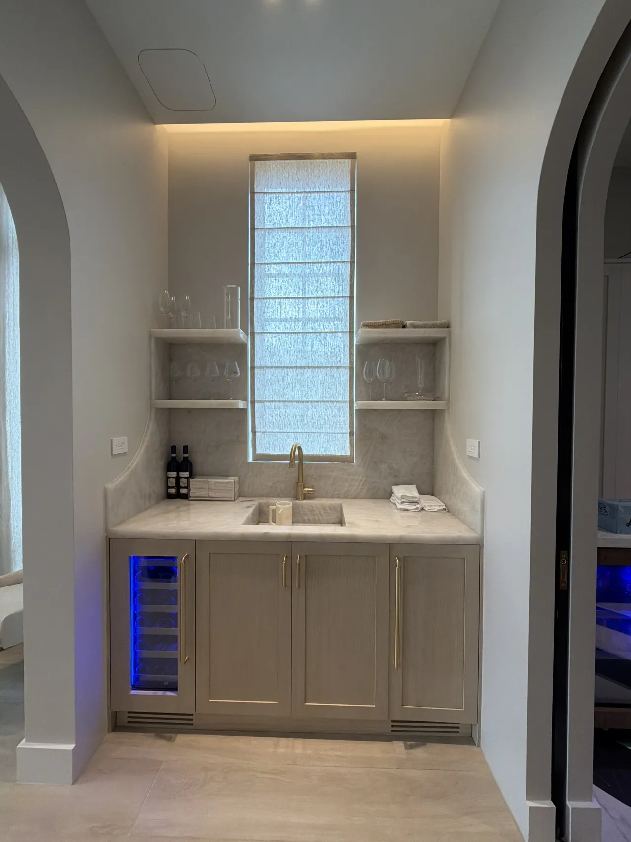 Wet bar alcove with a shaded window, stone counter and backsplash, corner shelves with glassware, taupe cabinets, and a blue-lit wine fridge