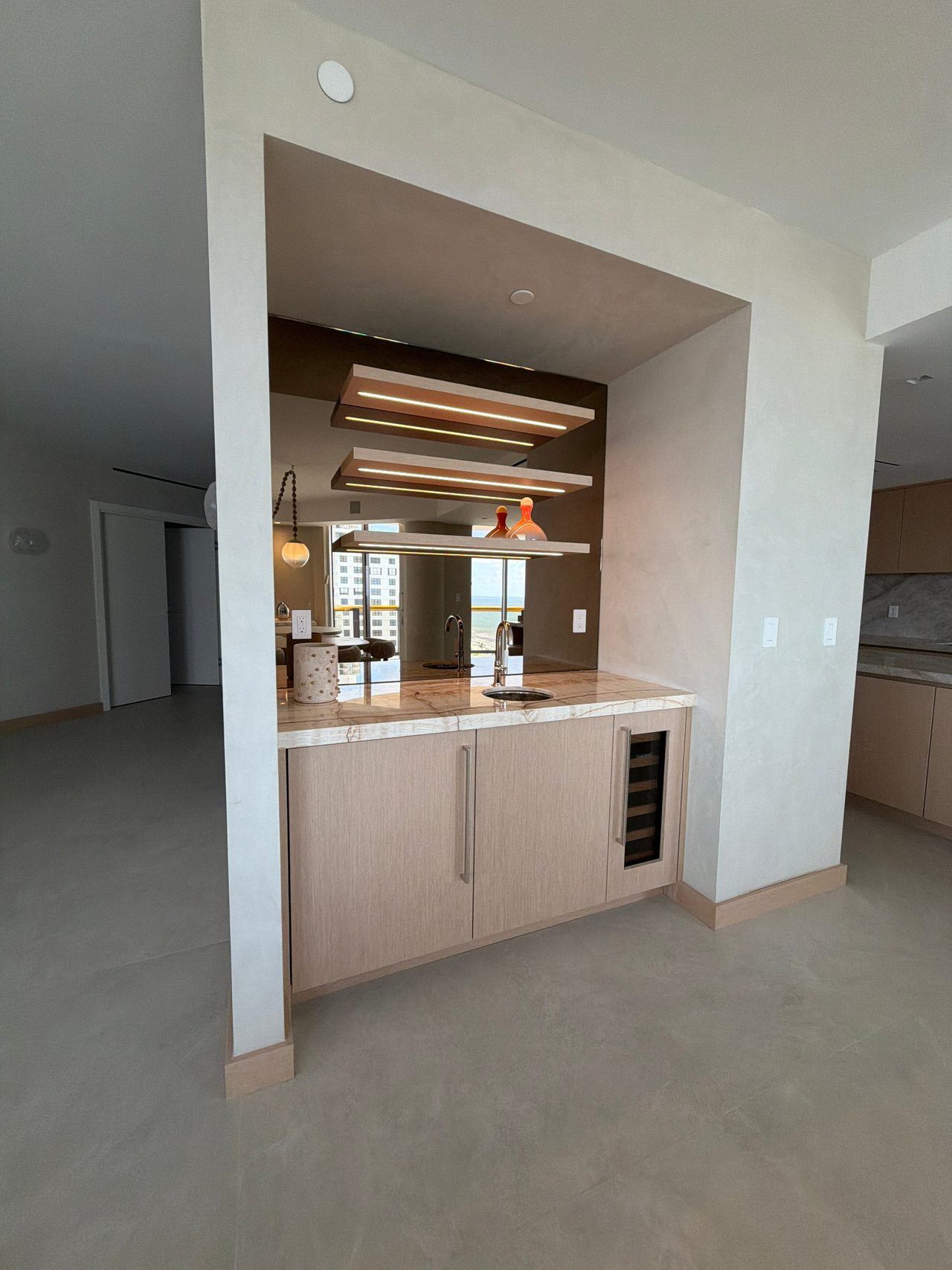 Wet bar alcove in custom-stained wood veneer, with integrated-LED floating shelves, a bronze mirror back panel, veined stone counter and wine refrigerator