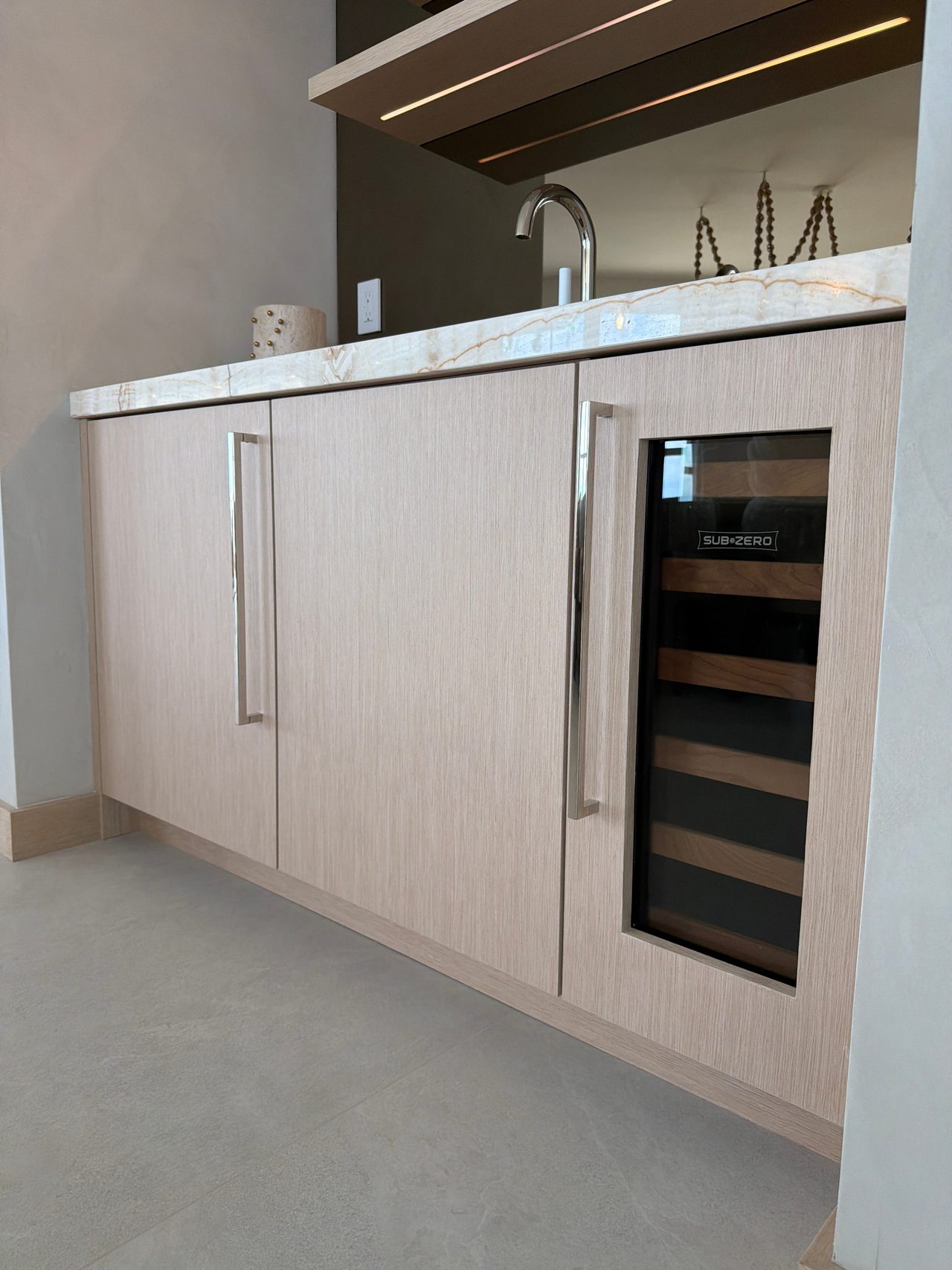Custom-stained wood veneer wet bar cabinets with chrome pulls and a glass-front wine refrigerator, veined stone counter and integrated-LED shelves above