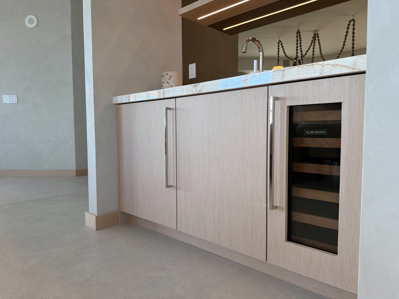 Custom-stained wood veneer wet bar cabinets with long chrome pulls, a veined-stone counter, and a glass-front undercounter wine refrigerator