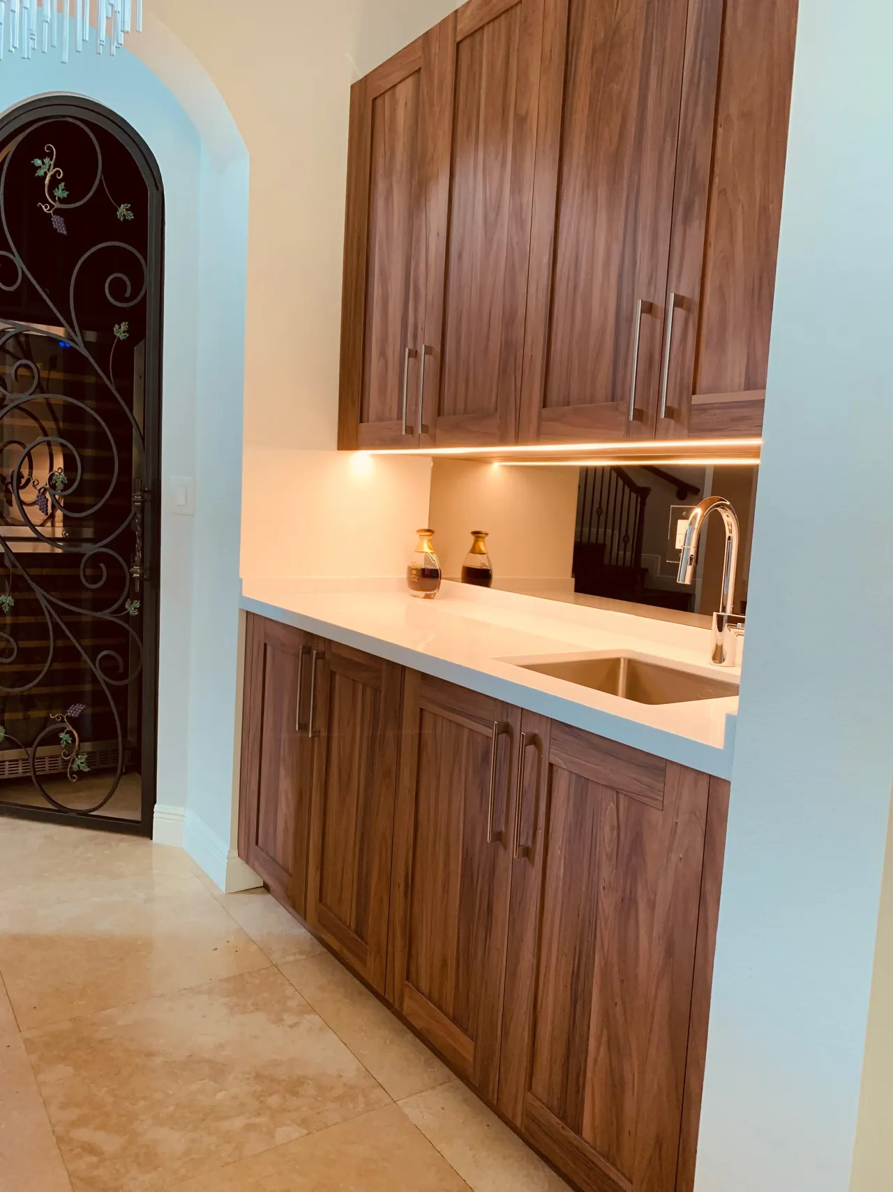 Wet bar with dark wood-grain cabinets, a white stone counter, an undermount sink with chrome faucet, a mirrored backsplash and an arched iron gate