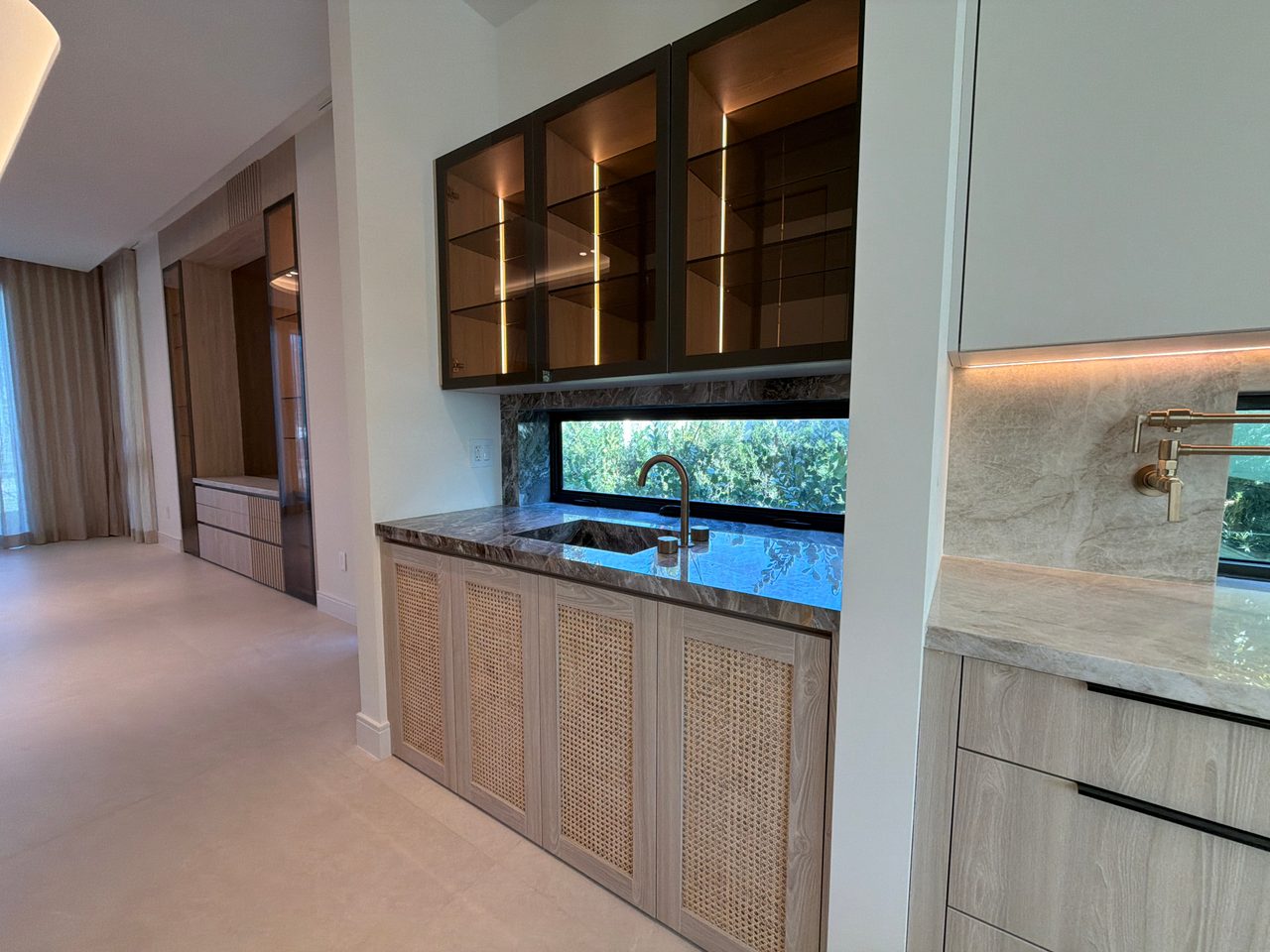 Wet bar nook with cane-front lower cabinets, a dark veined-stone counter, a ribbon window over the sink, and black-framed lit glass upper cabinets