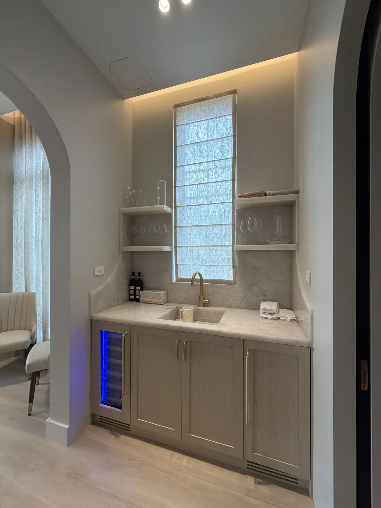 Wet bar nook framed by arched openings, with taupe cabinets, a stone counter and backsplash, shelves of glassware, and a wine fridge