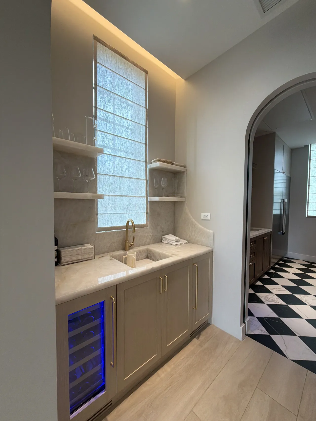 Wet bar with taupe shaker cabinets, a light stone counter and shelves, a gold-tone faucet, a blue-lit wine fridge, and a tall shaded window