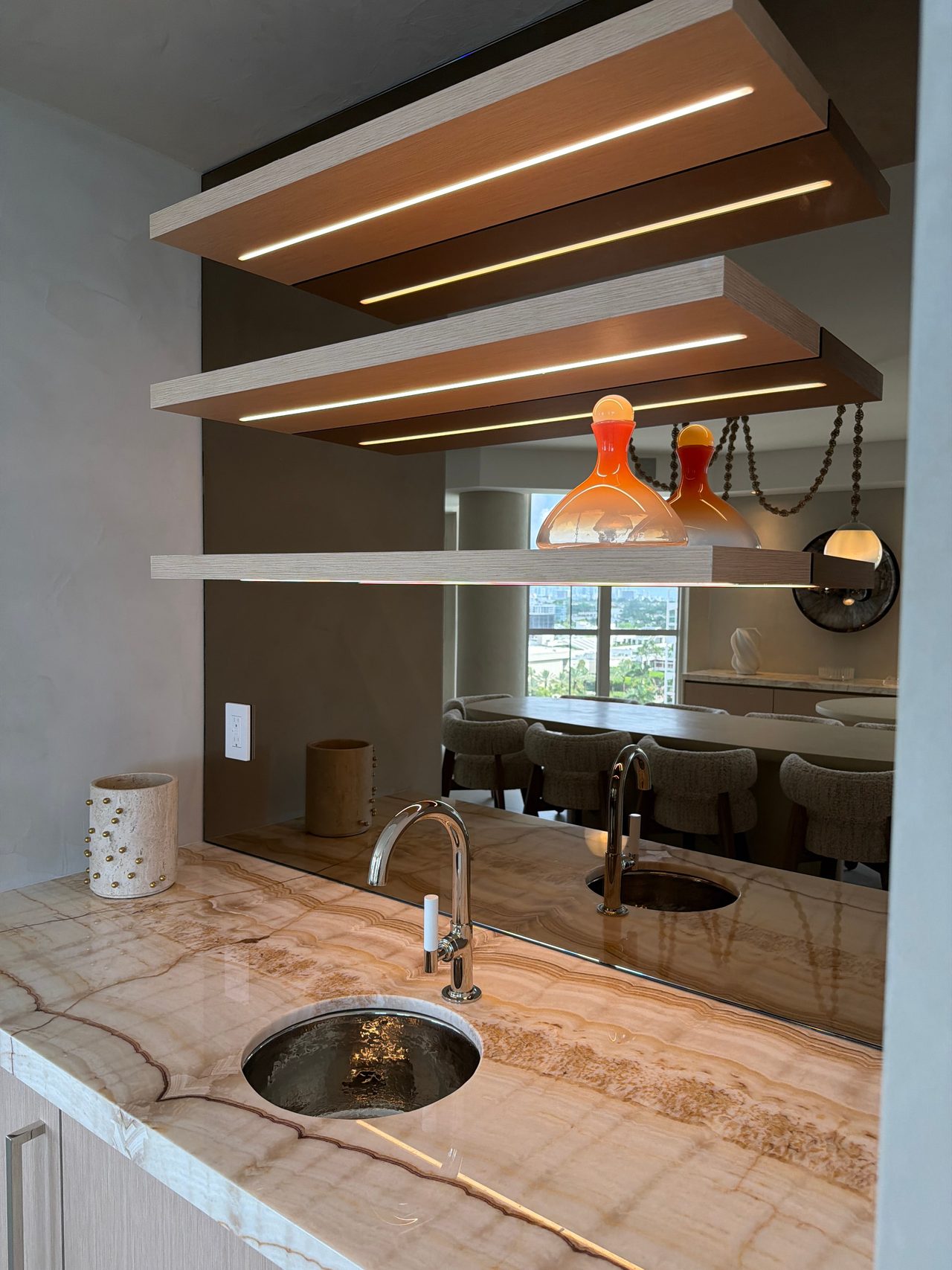 Veined stone wet bar counter with a hammered undermount sink and chrome faucet, under three custom-stained wood veneer shelves lit by integrated LED strips