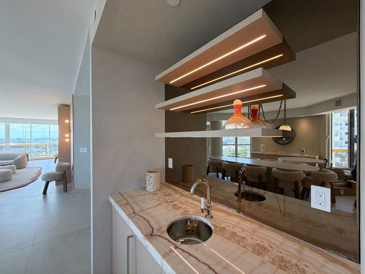 Veined-stone wet bar counter with an undermount sink and chrome faucet, below three integrated-LED custom-stained wood veneer shelves and a bronze mirror