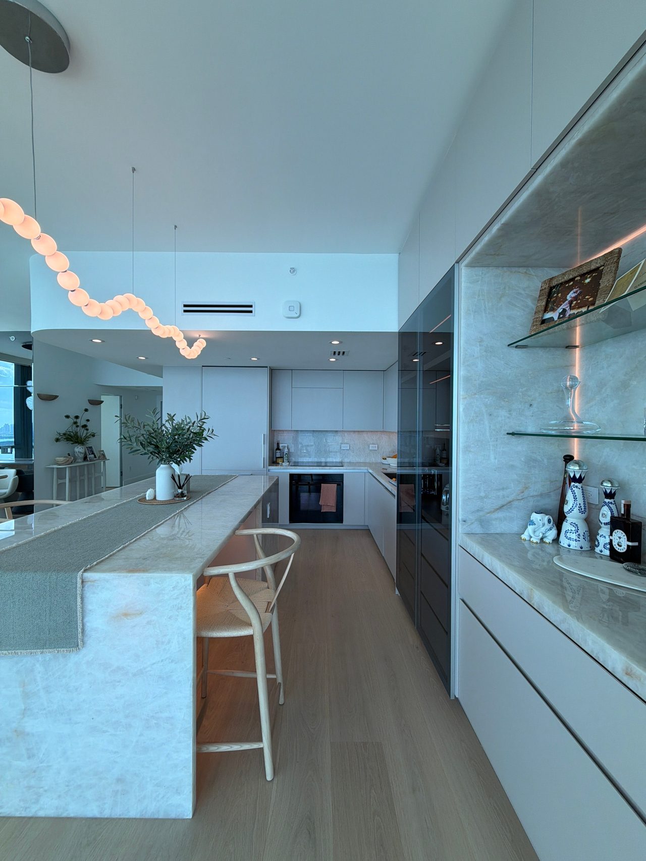 Kitchen with white flat-panel cabinetry, a built-in oven, a veined-stone island with a woven-seat stool, and a lit display niche with glass shelves